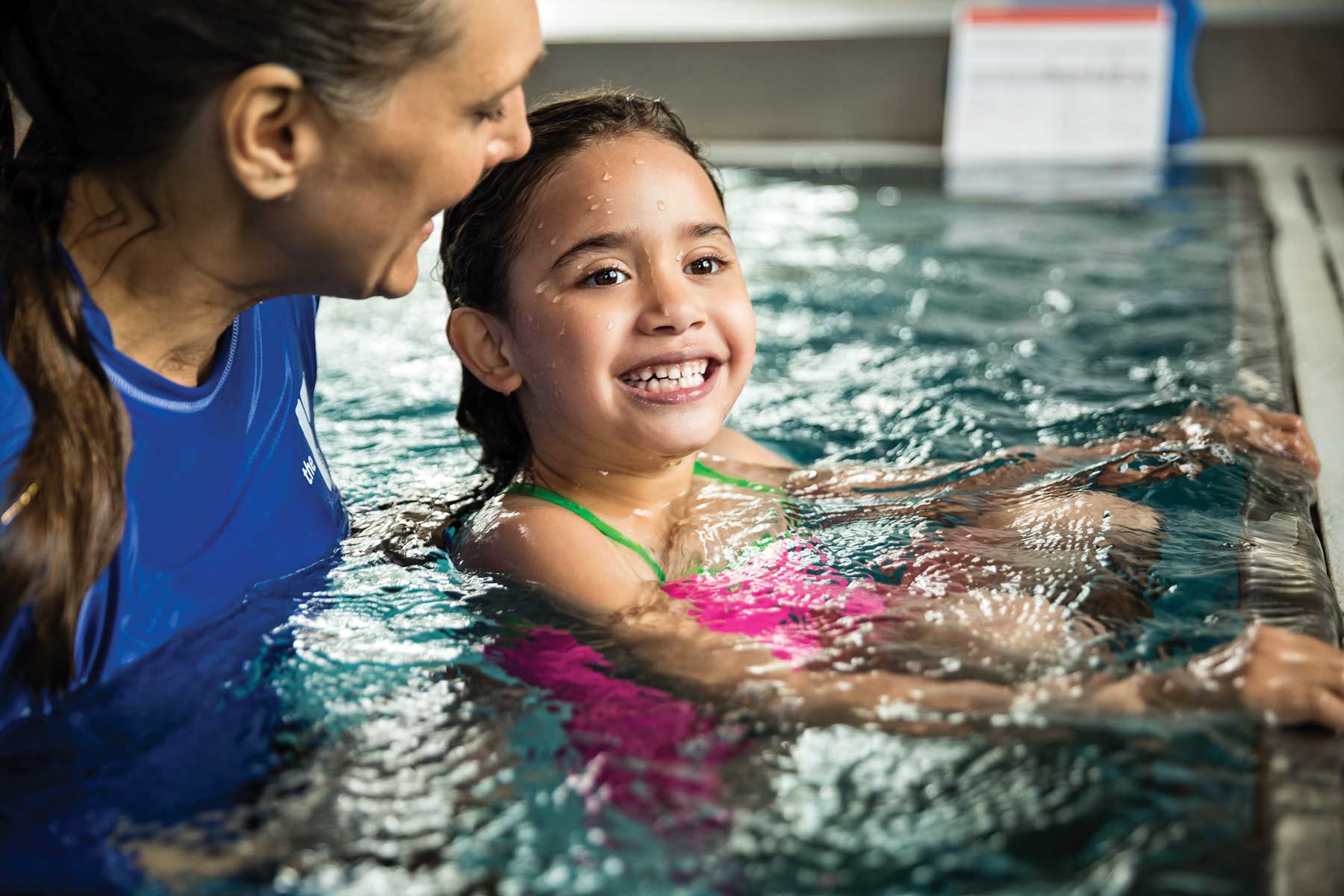 A girl smiles in the pool while learning to swim during a YMCA swimming lesson.
