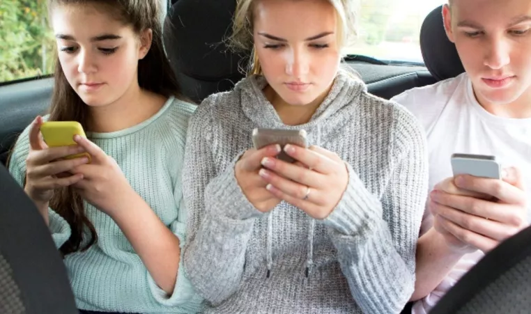 Three kids sitting in the backseat of a car with their cellphones